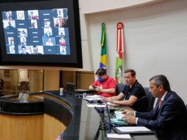 Alesc proíbe o corte de energia elétrica, água, esgoto e gás neste ano em SC julio garcia na mesa do parlamento com dois homens ao lado e deputados em transmissão no telão