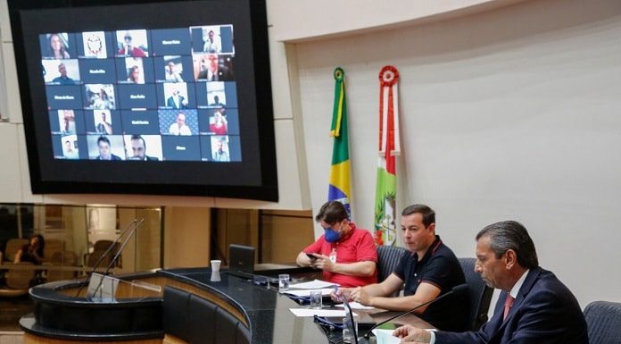 julio garcia na mesa do parlamento com dois homens ao lado e deputados em transmissão no telão