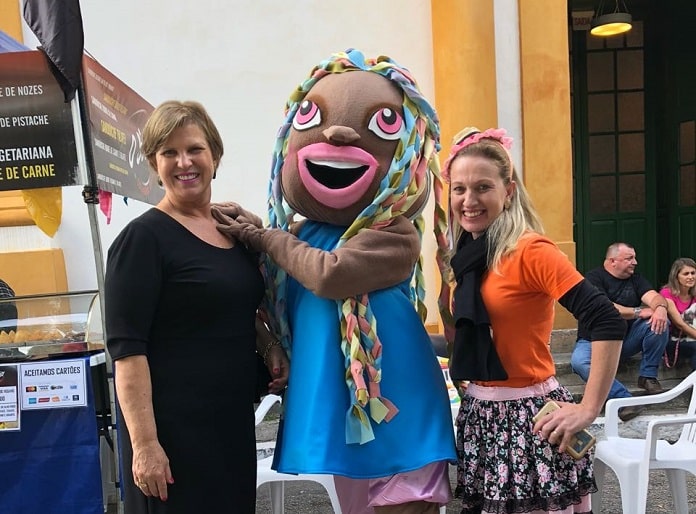 prefeita e secretária posam abraças para a foto com a mascote da feira