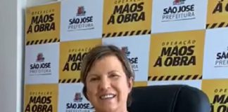 adeliana sentada atrás de mesa olha sorridente para câmera com painel da prefeitura ao fundo com logos