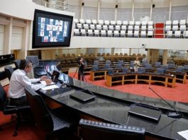 garcia sozinho na mesa diretora no plenário vazio da alesc com parlamentares em vídeos no telão
