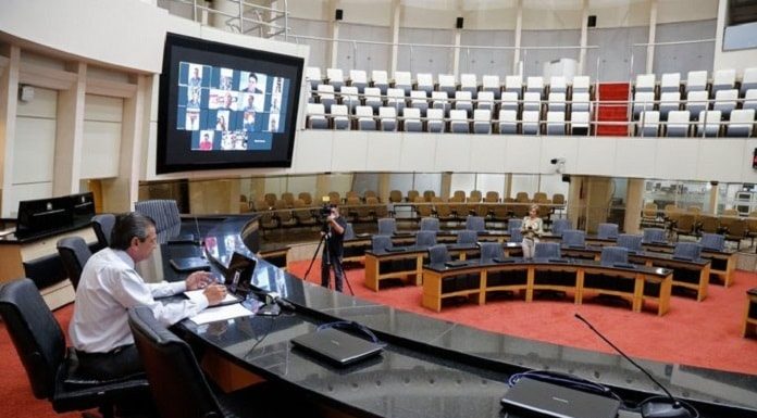 garcia sozinho na mesa diretora no plenário vazio da alesc com parlamentares em vídeos no telão