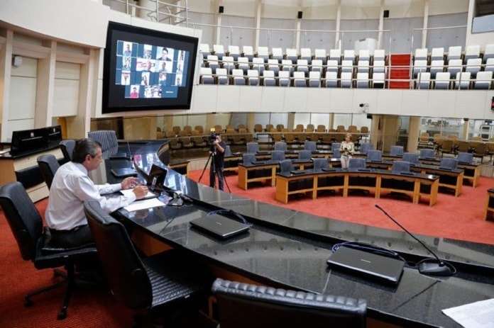 garcia sozinho na mesa diretora no plenário vazio da alesc com parlamentares em vídeos no telão