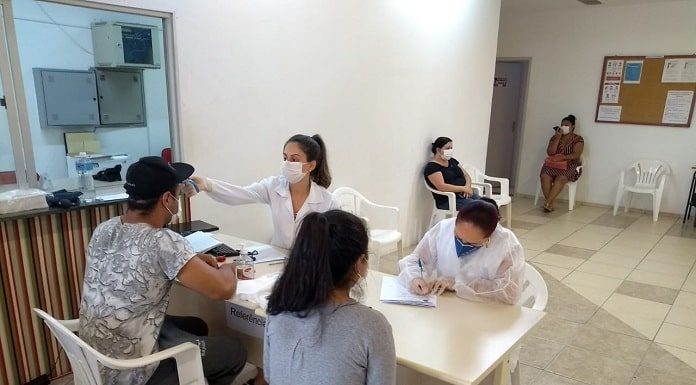 duas profissionais de saúde sentadas em mesas atendem duas pessoas; todos usam máscaras