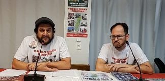 presidente e outro membro do sindicato com camisetas de campanha sentado atrás de mesa com panfletos e com microfones na frente