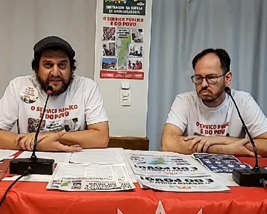 Sintrasem anuncia que não fará greve diante das restrições pela pandemia de Coronavírus presidente e outro membro do sindicato com camisetas de campanha sentado atrás de mesa com panfletos e com microfones na frente