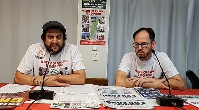 presidente e outro membro do sindicato com camisetas de campanha sentado atrás de mesa com panfletos e com microfones na frente