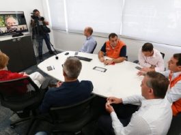 moisés e equipe reunidos ao redor de mesa olhando para uma tv onde está o ministro paulo guedes