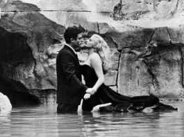 Mostra de filmes de Fellini celebra centenário do cineasta italiano cena do filme em que homem e mulher abraçados na água do mar próximo ao costão