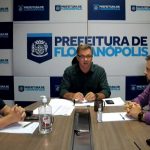 gean atrás de uma mesa com dois homens sentados de cada lado e painel da prefeitura ao fundo