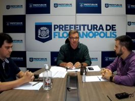 Prefeituras da Grande Florianópolis vão comprar testes de coronavírus em conjunto gean atrás de uma mesa com dois homens sentados de cada lado e painel da prefeitura ao fundo