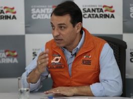 Processo de impeachment de Moisés é suspenso pelo Tribunal de Justiça moisés fala gesticulando sentado atrás de mesa usando colete da defesa civil e painel com logos do governo do estado ao fundo