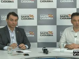 moisés e zeferino sentado atrás de mesa e no fundo painel do governo com logos