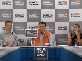 moisés e mais quatro pessoas em bancada de coletiva com painel ao fundo com logos do governo de sc