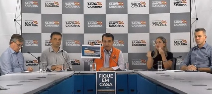 moisés e mais quatro pessoas em bancada de coletiva com painel ao fundo com logos do governo de sc