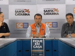moisés sentado atrás de mesa onde há um cartaz "fique em casa", secretário de saúde de um lado e intérprete de libras de outro