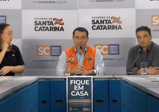 Governo estadual priorizará contratações para o quadro da saúde moisés sentado atrás de mesa onde há um cartaz "fique em casa", secretário de saúde de um lado e intérprete de libras de outro