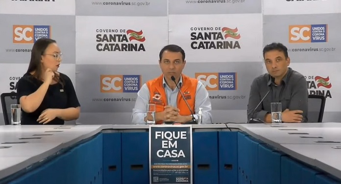 moisés sentado atrás de mesa onde há um cartaz "fique em casa", secretário de saúde de um lado e intérprete de libras de outro