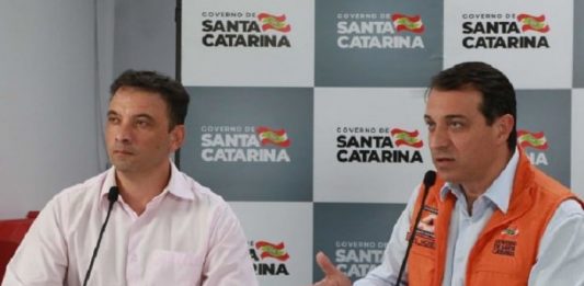 Com mais 713 leitos de UTI, Moisés planeja uma evolução controlada dos casos de Covid-19 em SC zeferino e moisés em bancada; moisés fala ao microfone; painel do governo com logos ao fundo