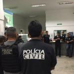 policiais civis reunidos em um hall conversando em pé; em primeiro plano agente de costas para foto com inscrição "polícia civil" nas costas