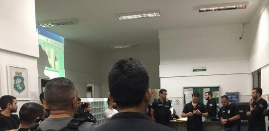 policiais civis reunidos em um hall conversando em pé; em primeiro plano agente de costas para foto com inscrição "polícia civil" nas costas