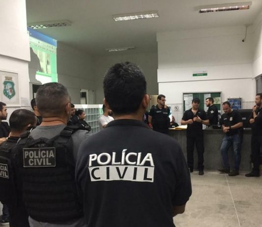 policiais civis reunidos em um hall conversando em pé; em primeiro plano agente de costas para foto com inscrição "polícia civil" nas costas