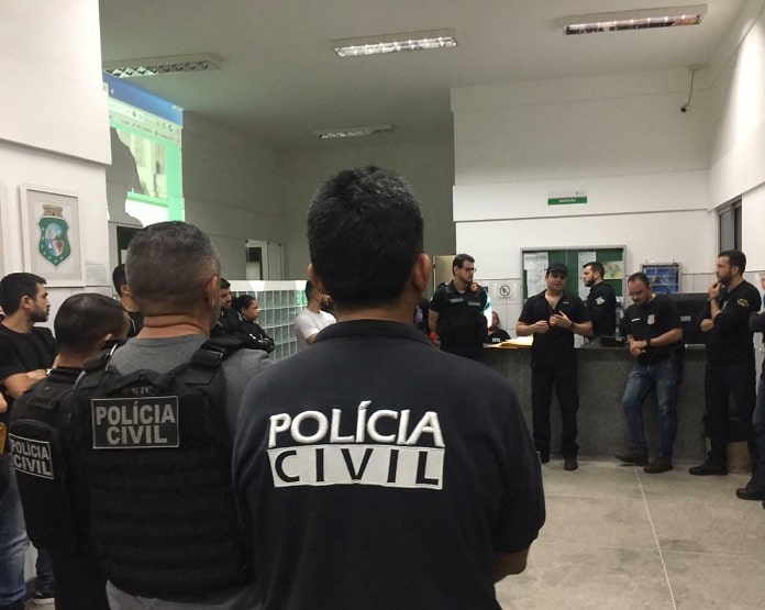 policiais civis reunidos em um hall conversando em pé; em primeiro plano agente de costas para foto com inscrição 