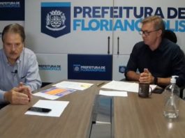 secretário e prefeito sentados na ponta de mesa com painel ao fundo com logos da prefeitura