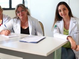 três médicas, duas de meia idade e outra mais jovem, sentadas atrás de mesa de consultório olham para a câmera