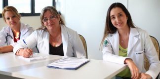 três médicas, duas de meia idade e outra mais jovem, sentadas atrás de mesa de consultório olham para a câmera