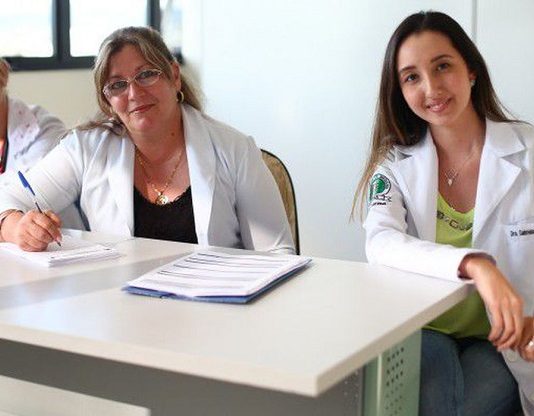 três médicas, duas de meia idade e outra mais jovem, sentadas atrás de mesa de consultório olham para a câmera