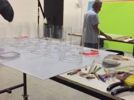 peças de protetores faciais sobre mesa em laboratório com ferramentas espalhadas e homem de costas ao fundo