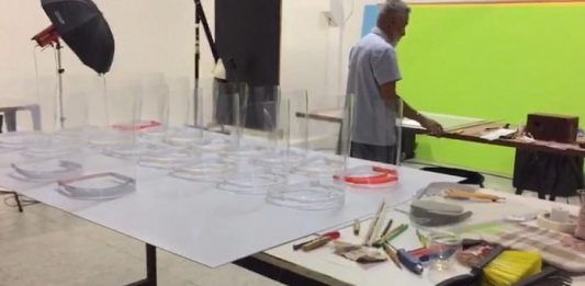 peças de protetores faciais sobre mesa em laboratório com ferramentas espalhadas e homem de costas ao fundo