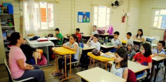 crianças sentadas em carteiras em sala de aula de madeira com professora à frente