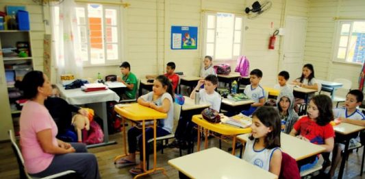 Educação da capital promove seminário de estágio, pesquisa e extensão crianças sentadas em carteiras em sala de aula de madeira com professora à frente