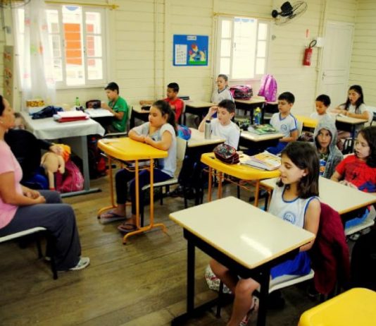 Educação da capital promove seminário de estágio, pesquisa e extensão crianças sentadas em carteiras em sala de aula de madeira com professora à frente