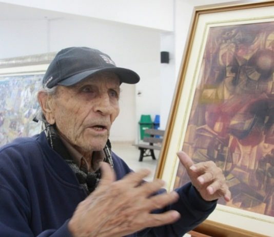 Sílvio Pléticos falece aos 95 anos pléticos usando boné gesticula conversando com um quadro seu ao lado