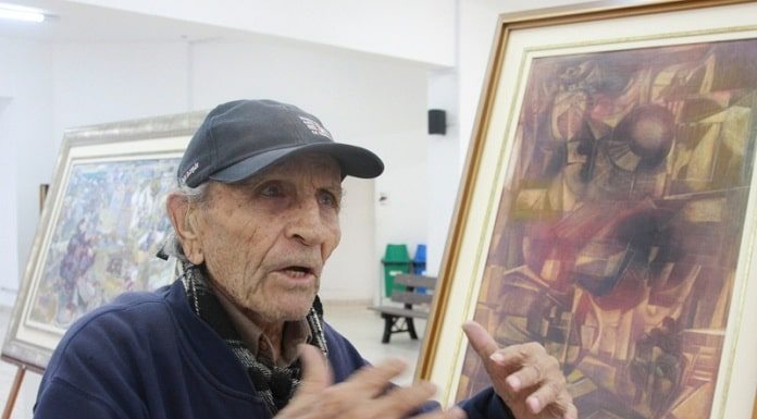 pléticos usando boné gesticula conversando com um quadro seu ao lado