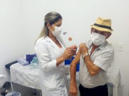 idoso com máscara no rosto usando chapéu de palha recebe vacina no braço esquerdo de uma profissional de saúde também usando máscara