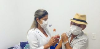 idoso com máscara no rosto usando chapéu de palha recebe vacina no braço esquerdo de uma profissional de saúde também usando máscara
