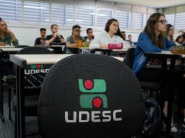carteira vazia com logo da udesc bordado e alunos na sala de aula em volta