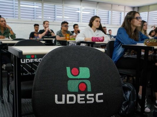 carteira vazia com logo da udesc bordado e alunos na sala de aula em volta