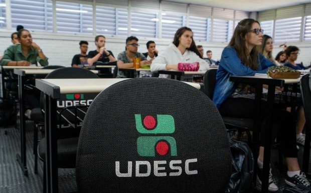 carteira vazia com logo da udesc bordado e alunos na sala de aula em volta