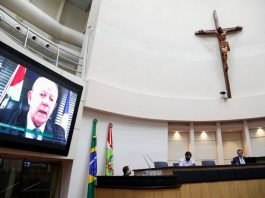 Alesc aprova que atividades religiosas e físicas são essenciais em calamidade pública grande cruz sobre a mesa diretora da alesc, com deputado em transmissão no telão