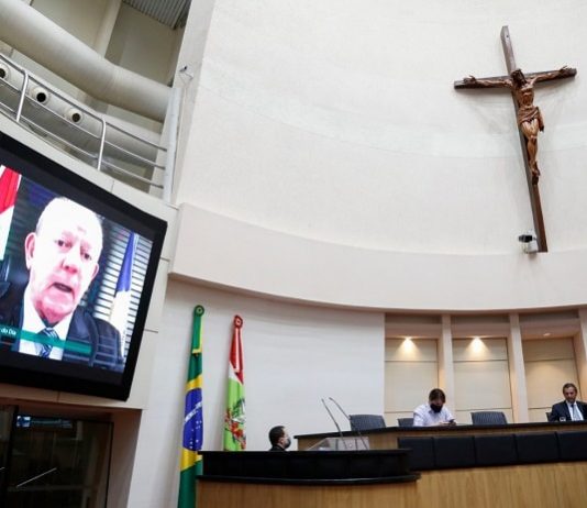 grande cruz sobre a mesa diretora da alesc, com deputado em transmissão no telão