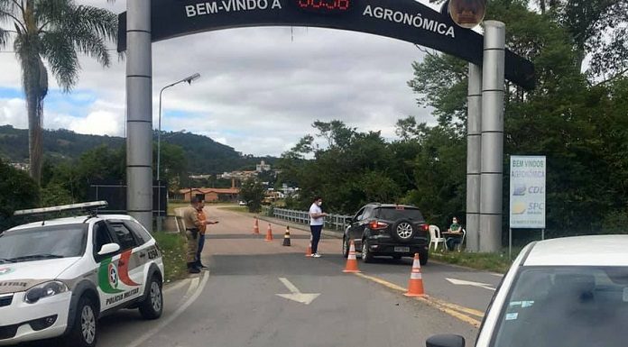 carro parado no sob o portal da cidade onde há cones na estrada e um homem de máscara ao lado do carro; carro da polícia; mais três pessoas em volta