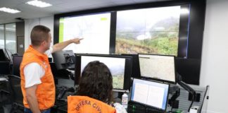 homem e mulher e mulher com coletes da defesa civil olhando para diversos monitores, em um deles há um rio seco