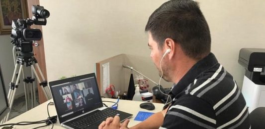 Câmara de SJ aprova criação de fundo para doações contra pandemia vereador michel schlemper sentado com notebook sobre mesa com transmissão dos outros vereadores e câmera de vídeo na frente da mesa