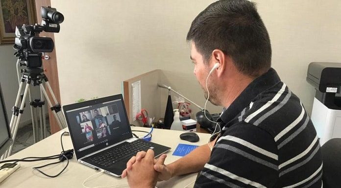 vereador michel schlemper sentado com notebook sobre mesa com transmissão dos outros vereadores e câmera de vídeo na frente da mesa