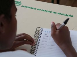 aluno escrevendo com caneta em caderno sobre carteira onde está escrito "secretaria estadual de educação"